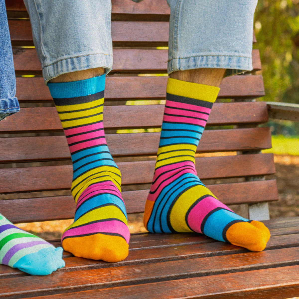 Two people sitting on a wooden park bench wearing colorful striped socks. One person wears green, purple, and blue striped socks, while the other wears bright multicolor striped socks in blue, pink, yellow, and orange.