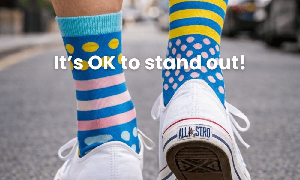 Colorful socks with polka dots and stripes worn on white sneakers, with text 'It's OK to stand out!' on a blurred street background.