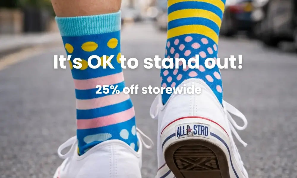Colorful socks with text overlay 'It's OK to stand out! 25% off storewide' on a blurred street background.